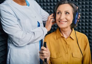 Portrait senior woman with white toothy smile getting hearing test at soundproof audiometric booth using audiometry headphones and audiometer